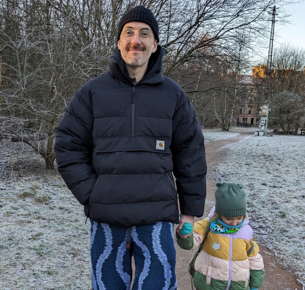 David white tall man that is using a big black jacket and a black hat, smiling holding a hand of a small kid
